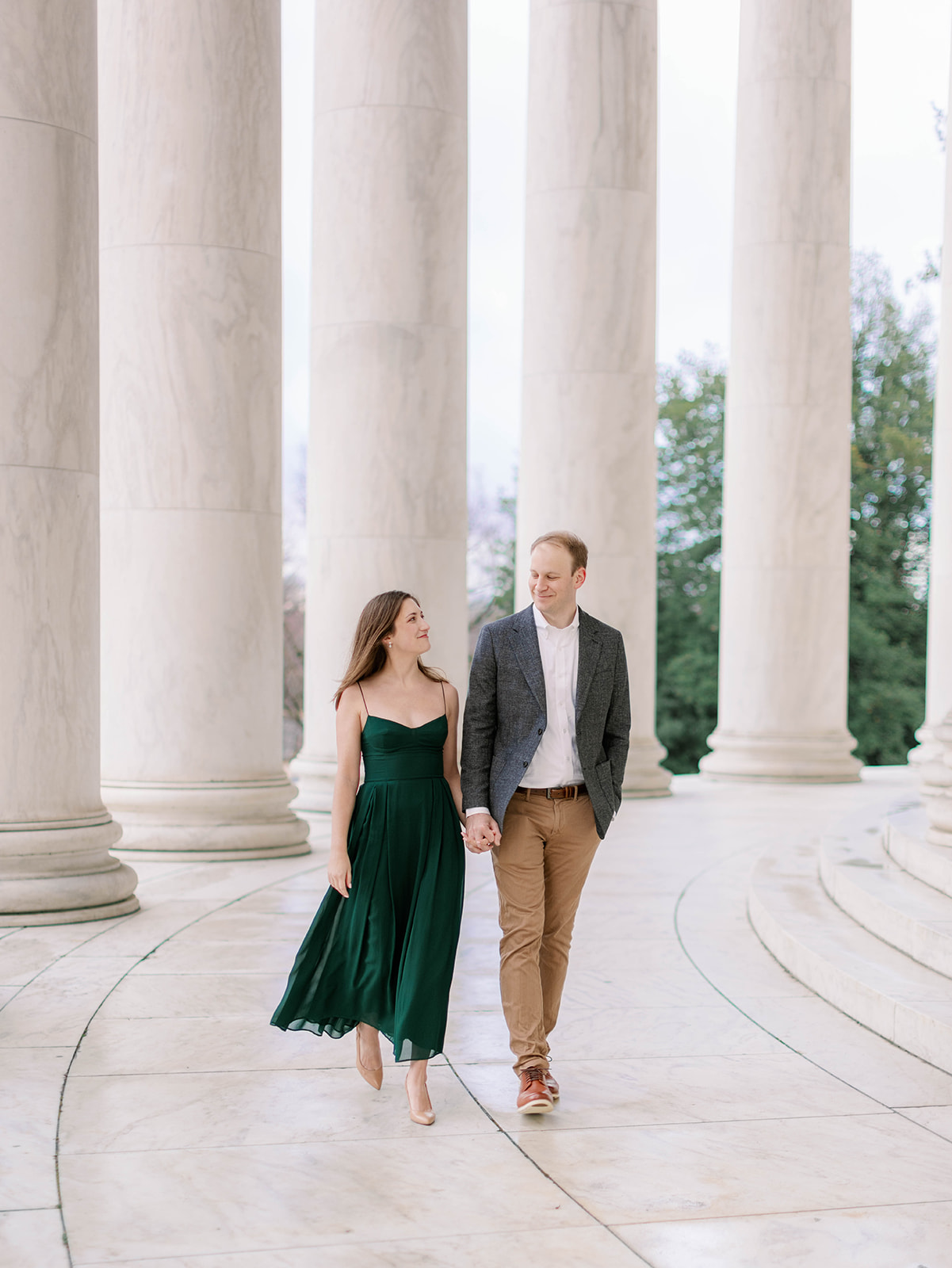 Couple walking among the columns at the Jefferson Memorial, one of the best engagement photo locations in DC. The woman is wearing a green dress and the man is wearing brown chinos, brown shoes and a grey jacket.