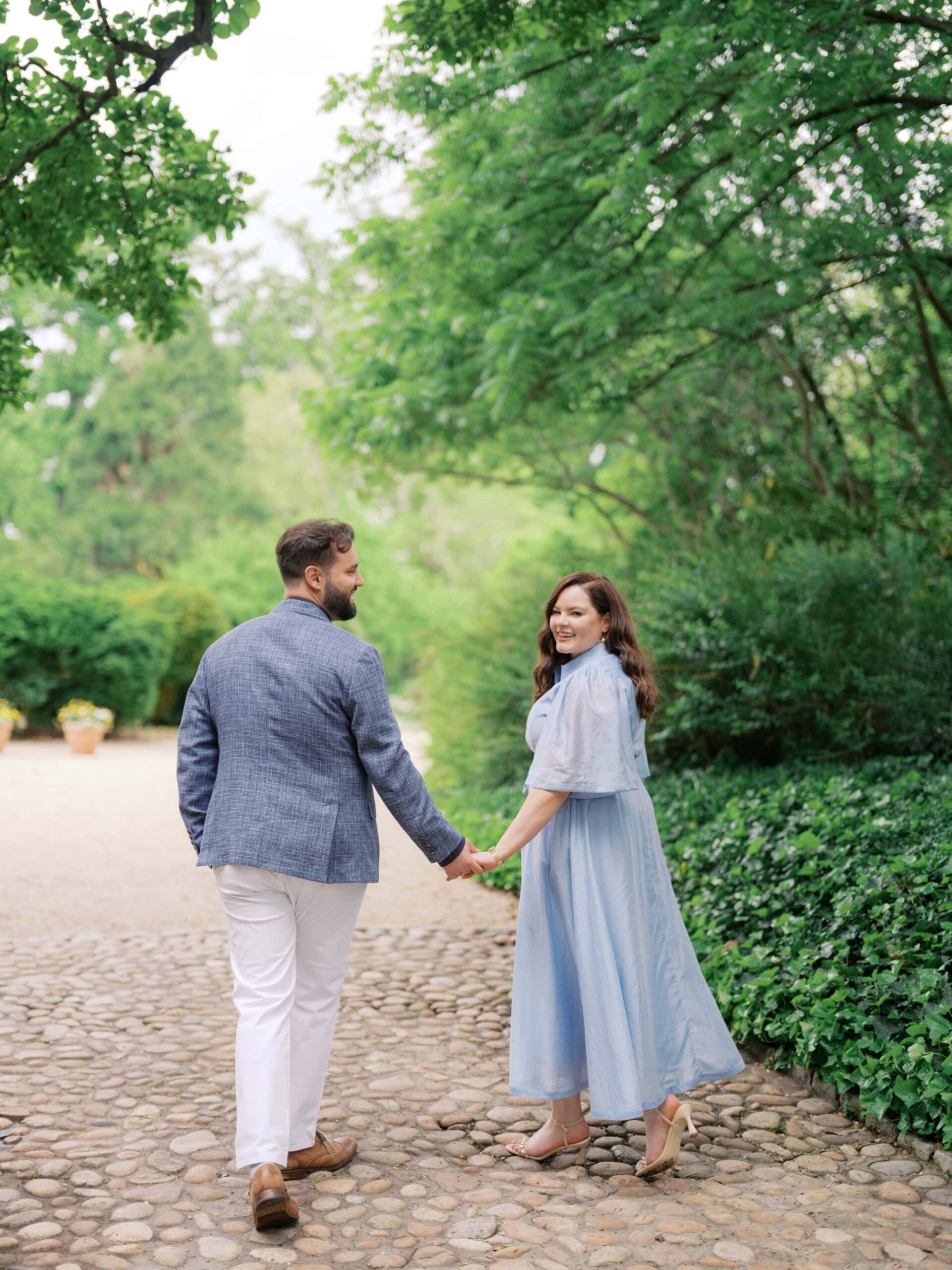 Couple walking at Tudor Place, a historic home and garden in Georgetown, one of the best engagement photo locations in DC. The woman is wearing a blue dress and the man is wearing white chinos, brown shoes and a blue jacket.