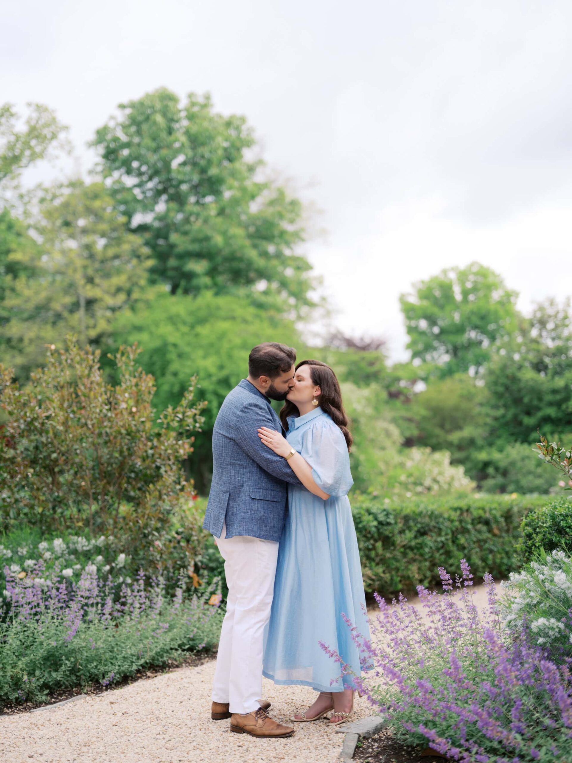 Couple kissing at Tudor Place, a historic home and garden in Georgetown, one of the best engagement photo locations in DC. The woman is wearing a blue dress and the man is wearing white chinos, brown shoes and a blue jacket.