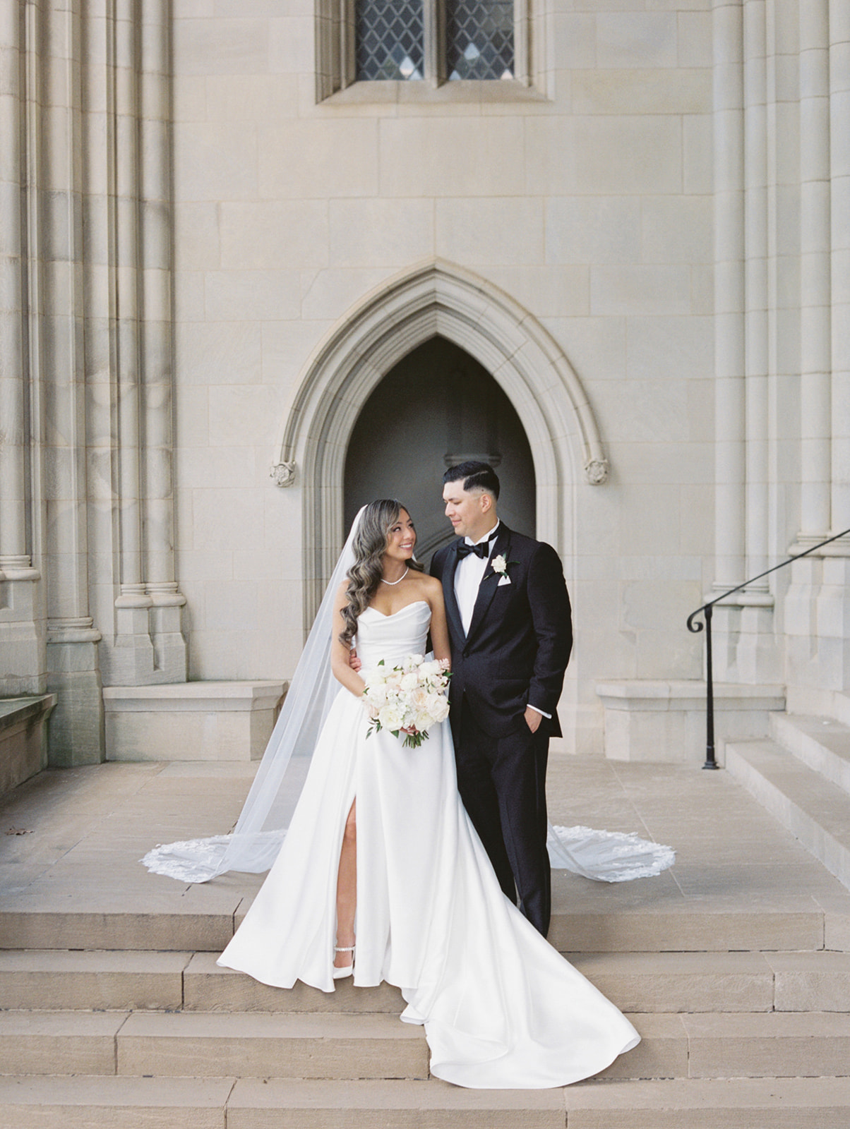 A bride and groom standing on the steps of the Washington National Cathedral, smiling at each other. The bride wears a white wedding dress and holds a bouquet of flowers, while the groom is in a black tuxedo.