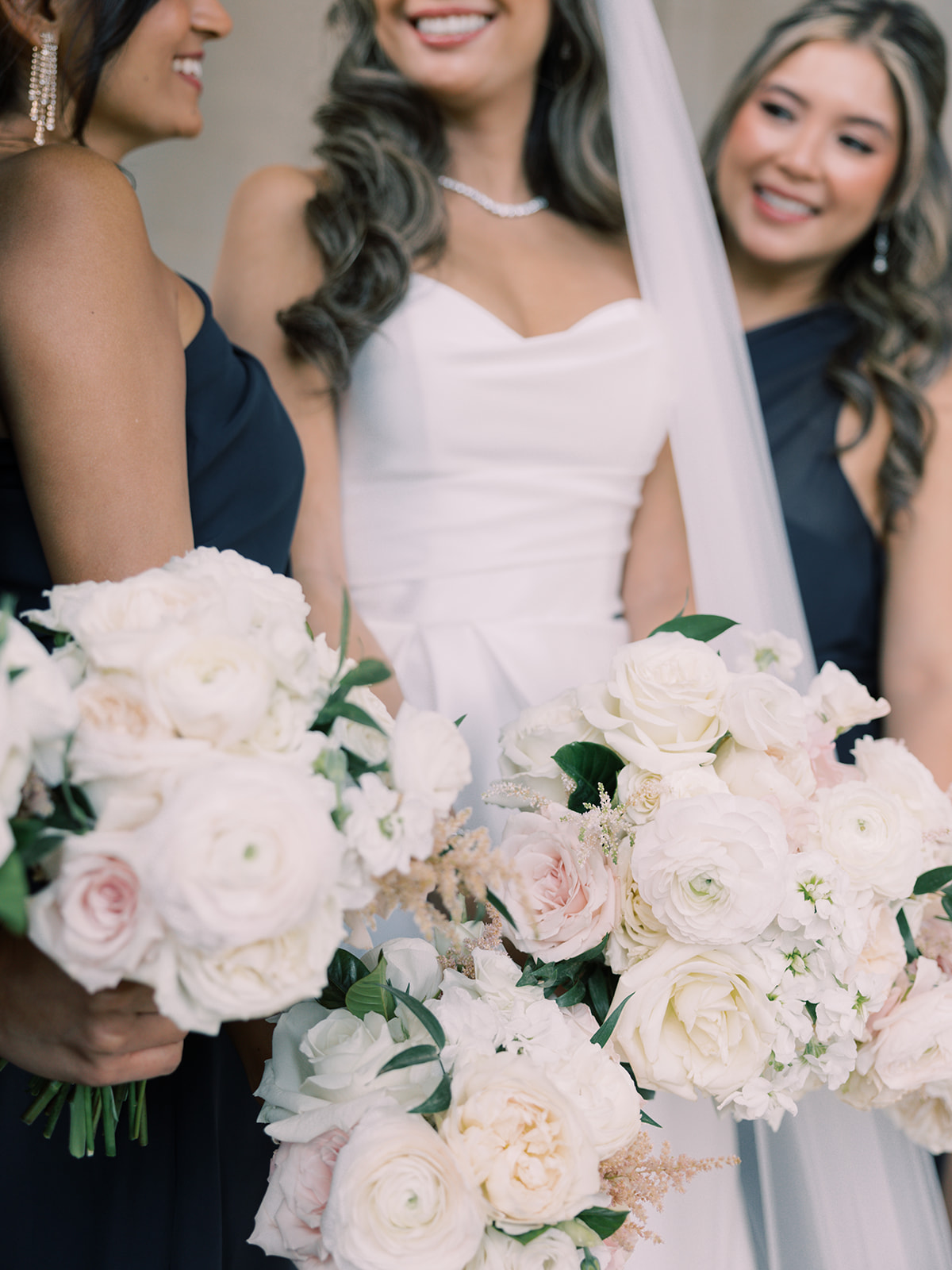 A close-up image of three women with floral bouquets, showcasing the bride in a white dress and her bridesmaids in dark dresses, all smiling.