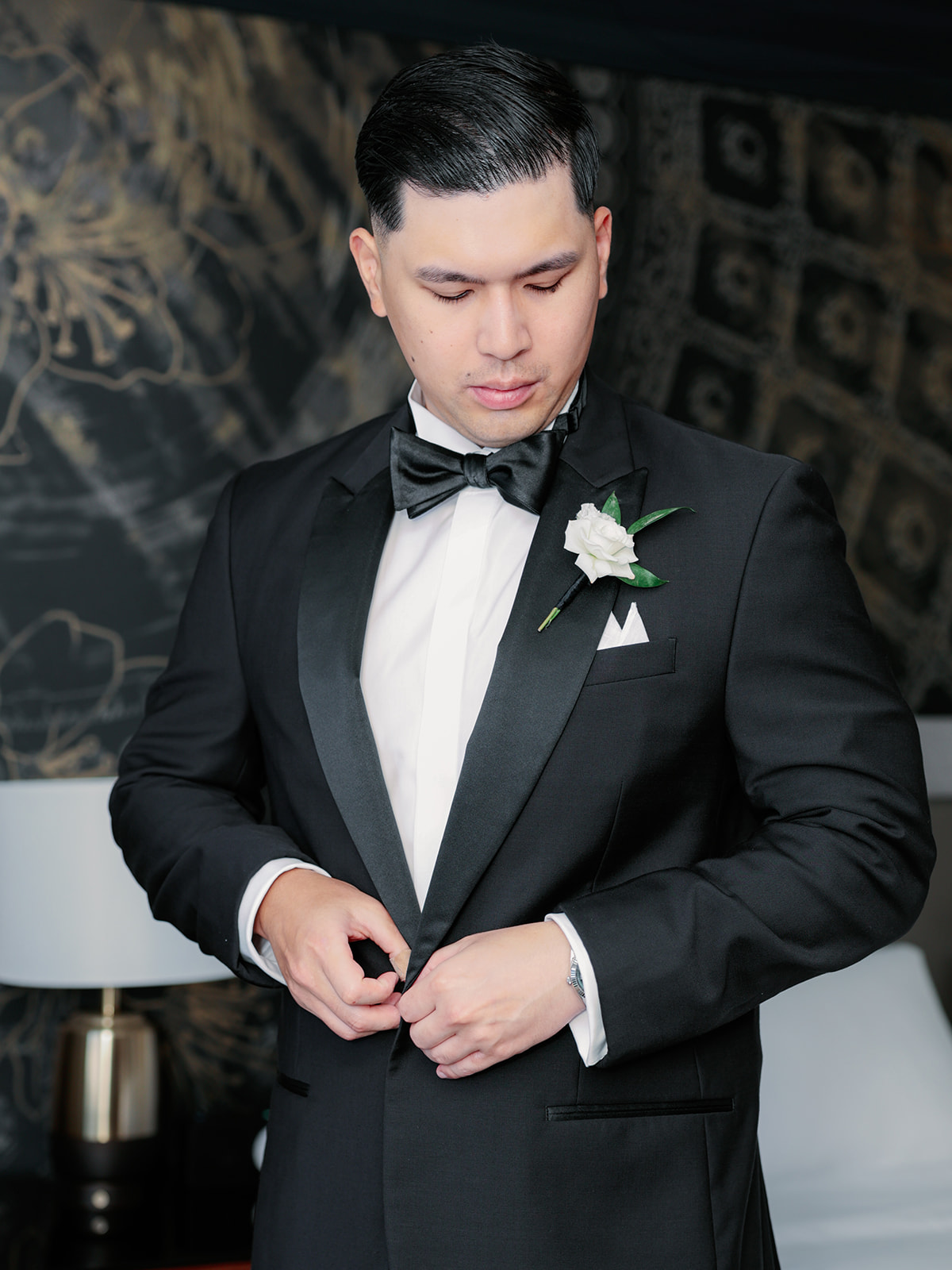 Groom adjusting his tuxedo jacket while wearing a bow tie and white rose boutonniere, with a dark patterned background.