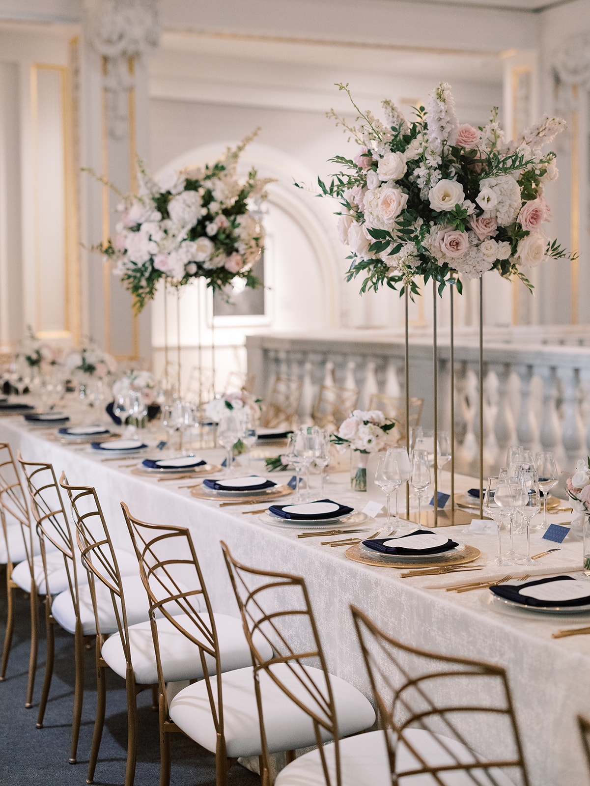 Elegant wedding reception at the National Museum of Women in the Arts, setup featuring tall floral arrangements, a long dining table adorned with gold accents, navy plates, and glassware in a beautifully decorated venue.