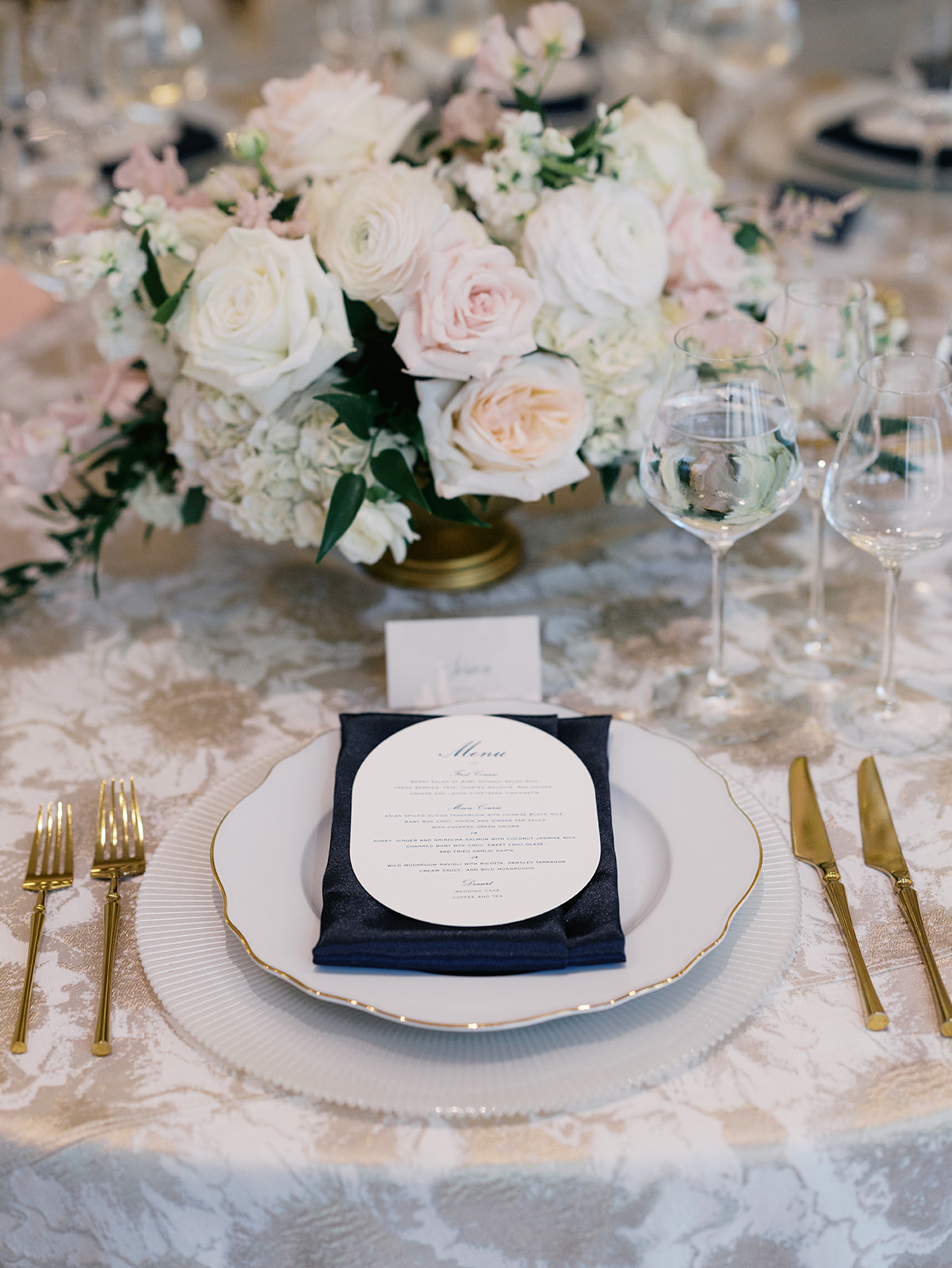 Elegant wedding table setting featuring a floral arrangement of white and pink roses, a navy napkin, and a menu card on a decorative plate. From a wedding in Washington DC at the National Museum of Women in the Arts.